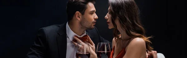 Panoramic shot of attractive woman adjusting bow tie of elegant man by wine glasses isolated on black — Stock Photo