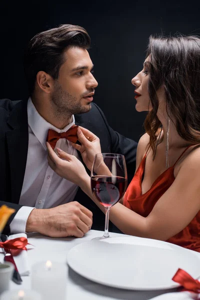 Mujer elegante ajustando corbata de lazo de hombre guapo durante la cena romántica aislado en negro - foto de stock