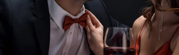 Обрезанный вид женщины регулируя бабочку галстук мужчины с бокалом вина изолированы на черном, панорамный снимок — стоковое фото