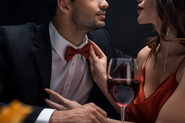 Vista recortada de mujer elegante ajustando corbata de lazo de hombre en traje por copa de vino aislado en negro - foto de stock