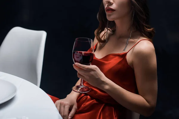 Vista ritagliata di donna elegante in abito rosso che tiene bicchiere di vino a tavola isolato su nero — Foto stock