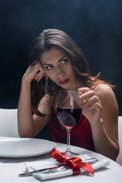 Attractive woman with hand by head touching wine glass at served table on black background with smoke — Stock Photo