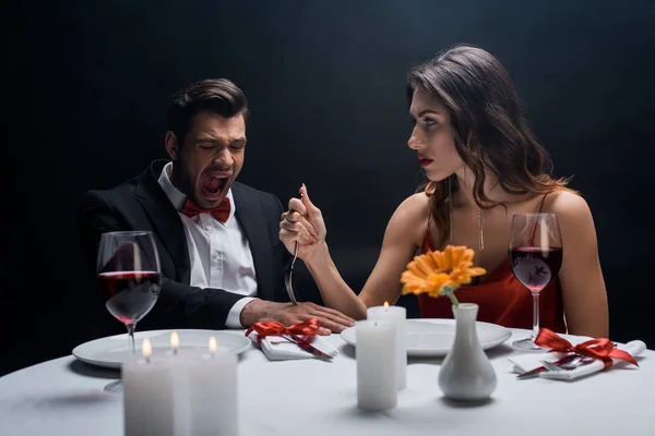 Hermosa mujer clavando tenedor en la mano de hombre gritando durante la cena romántica aislado en negro — Stock Photo