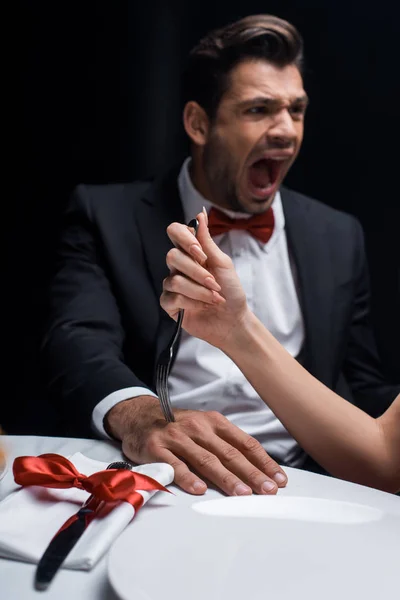 Selective focus of woman sticking fork in hand of screaming boyfriend during romantic dinner isolated on black — Stock Photo