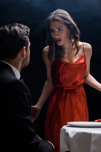 Mulher irritada olhando para o namorado por mesa servida no fundo preto com fumaça — Fotografia de Stock