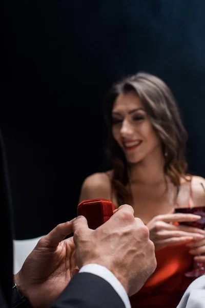 Focus selettivo dell'uomo che tiene la scatola con gioielli sorridendo donna con bicchiere di vino isolato su nero — Foto stock