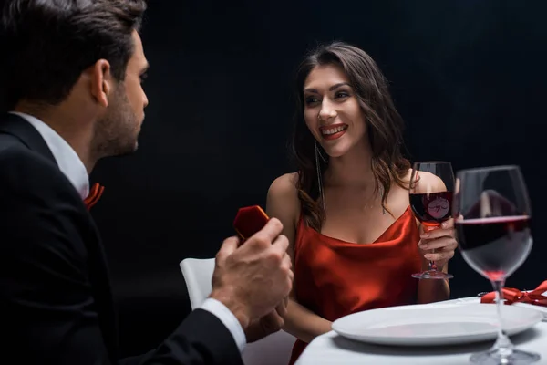Красивый мужчина представляет шкатулку с драгоценностями улыбающейся девушке во время романтического ужина, изолированного на черном — стоковое фото