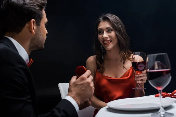 Handsome man presenting jewelry box at smiling girlfriend during romantic dinner isolated on black — Stock Photo