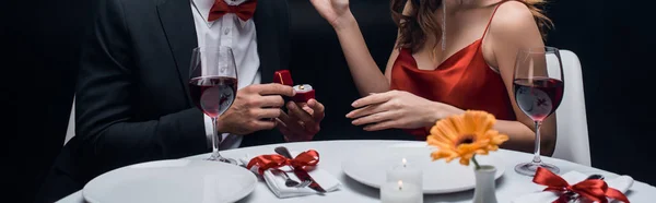Обрезанный вид мужчины, представляющего коробку с ювелирным кольцом на женщину во время романтического ужина, изолированного на черном, панорамный снимок — стоковое фото