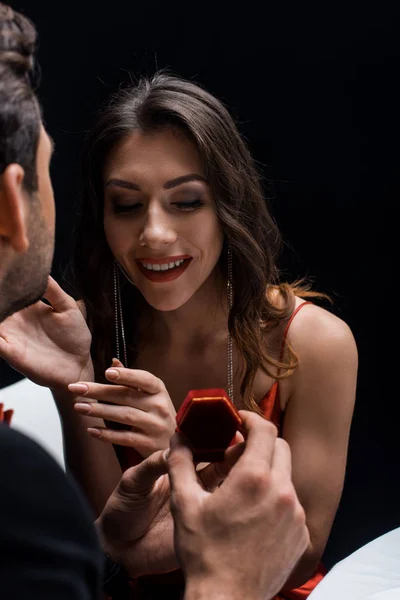Selektiver Fokus des Mannes präsentiert Schmuckschatulle bei lächelnder Freundin am Tisch isoliert auf Schwarz — Stockfoto