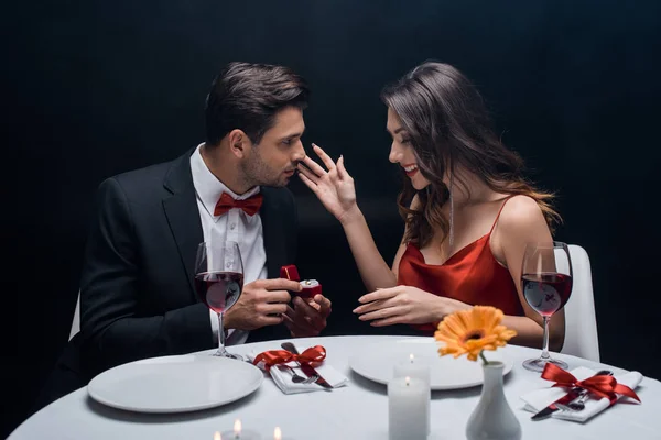 Side view of handsome man presenting jewelry ring at smiling girlfriend during romantic dinner isolated on black — Stock Photo