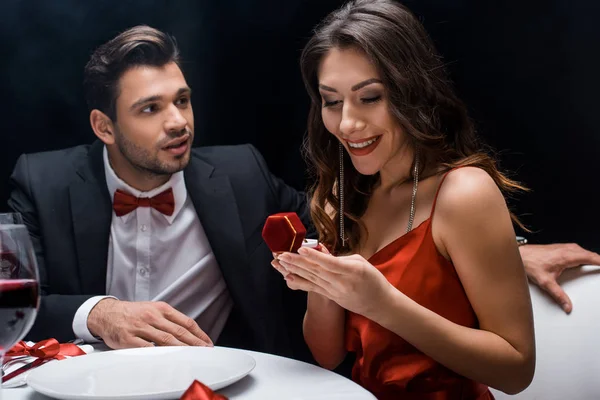 Beau homme regardant petite amie souriante avec bague à bijoux pendant le dîner romantique isolé sur noir — Photo de stock
