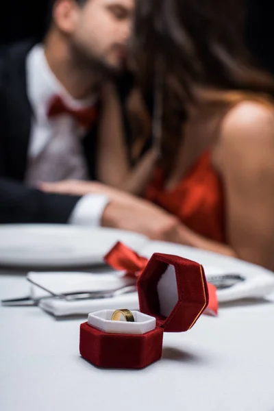 Selective focus of jewelry ring in box on served table with kissing couple at background isolated on black — Stock Photo