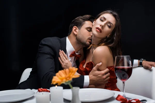 Handsome man kissing beautiful girlfriend during romantic dinner isolated on black — Stock Photo