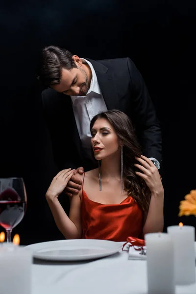 Enfoque selectivo del hombre guapo abrazando a la mujer elegante en la mesa servida aislada en negro - foto de stock