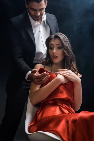 Low angle view of man presenting box with jewelry at shocked woman on black background with smoke — Stock Photo