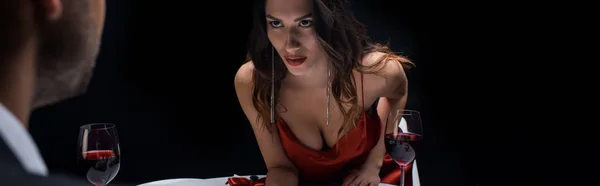 Foto panorámica de la hermosa mujer coqueteando con el hombre al lado de copas de vino en la mesa aislada en negro - foto de stock