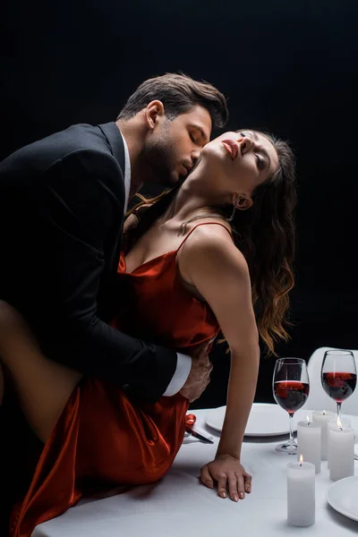 Vue latérale de bel homme embrassant belle femme dans le cou sur la table servie isolé sur noir — Photo de stock