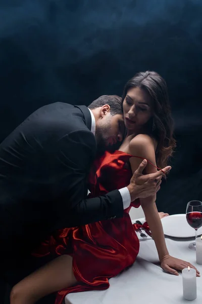 Vista lateral do homem bonito decolando vestido de mulher sexy durante o jantar romântico no fundo preto com fumaça — Fotografia de Stock