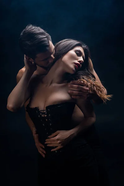 Handsome shirtless man kissing in neck attractive woman in corset isolated on black — Stock Photo