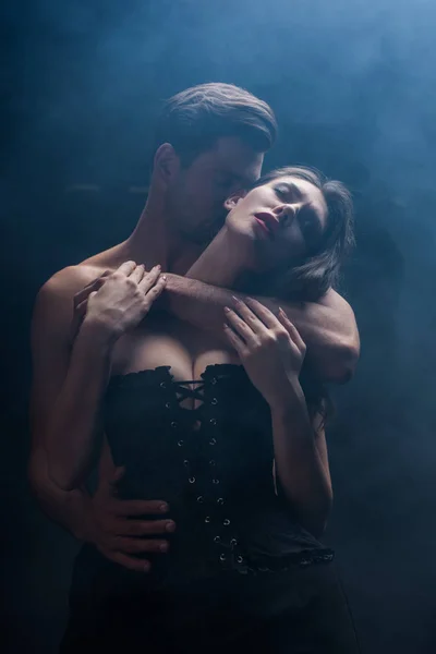 Hombre guapo besándose y abrazando a mujer seductora en corsé sobre fondo negro con humo - foto de stock