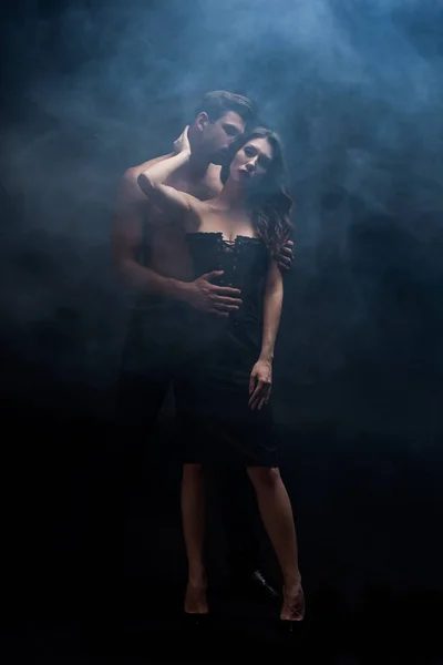 Повна довжина м'язистого чоловіка цілує сексуальну жінку на чорному тлі з димом — стокове фото