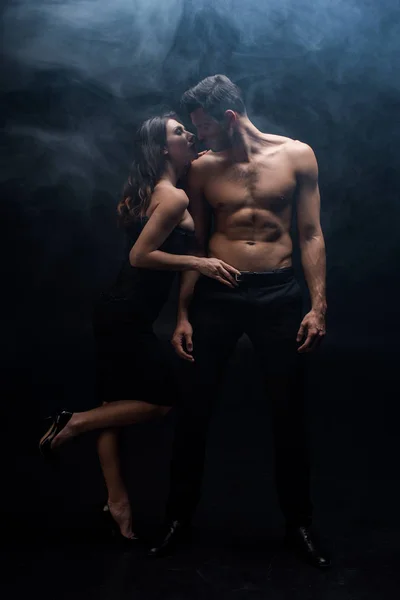 Pleine longueur de femme sensuelle touchant ceinture de l'homme musculaire sur fond noir avec de la fumée — Photo de stock