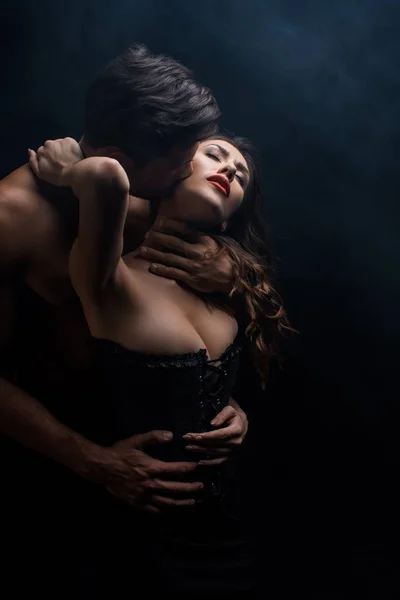 Hombre sin camisa besándose y abrazando a mujer sensual en corsé aislado en negro - foto de stock