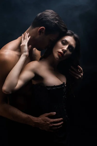 Muscular man touching sexy girlfriend in corset isolated on black — Stock Photo