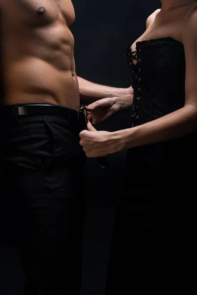 Cropped view of sexy woman in corset taking off belt of muscular man isolated on black — Stock Photo