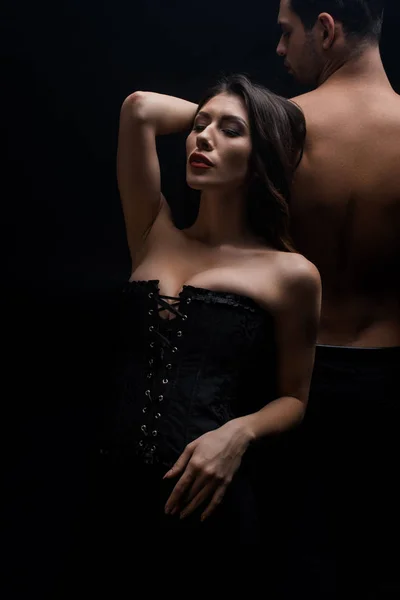 Donna sexy in corsetto in piedi da uomo senza camicia isolato su nero — Foto stock