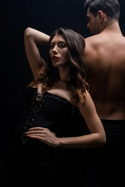 Femme sensuelle dans le corset debout à l'arrière du copain torse nu et regardant la caméra isolée sur noir — Photo de stock