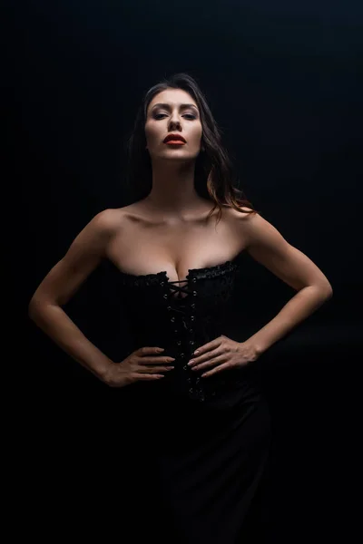 Femme sexy en corset avec la main sur les hanches en regardant la caméra isolée sur noir — Photo de stock