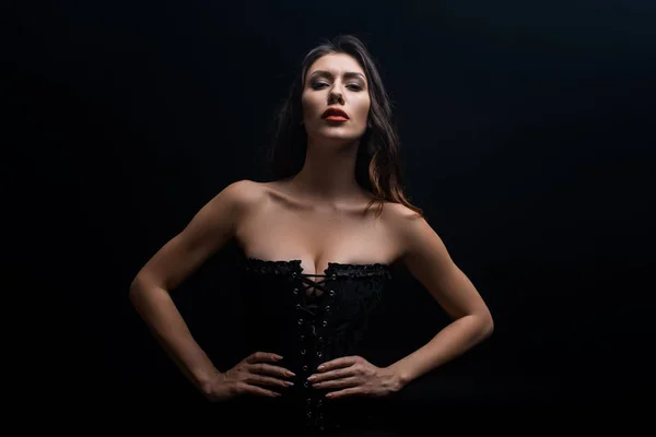 Belle femme en corset avec les mains sur les hanches en regardant la caméra isolée sur noir — Photo de stock