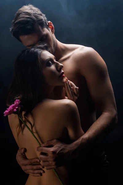 Handsome muscular man hugging beautiful naked girlfriend while holding gerbera on black background with smoke — Stock Photo
