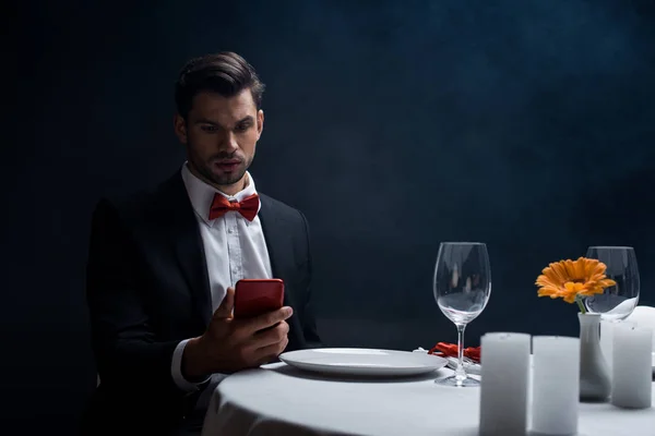 Elegante hombre mirando el teléfono inteligente en la mesa servida con velas sobre fondo negro con humo - foto de stock