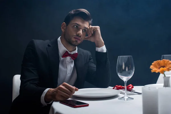 Elegante hombre con la mano por la cabeza sentado por teléfono inteligente en la mesa servida sobre fondo negro con humo - foto de stock