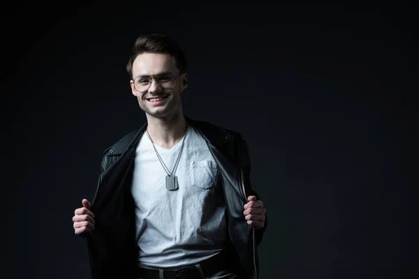 Smiling stylish brutal man with hands on biker jacket isolated on black — Stock Photo