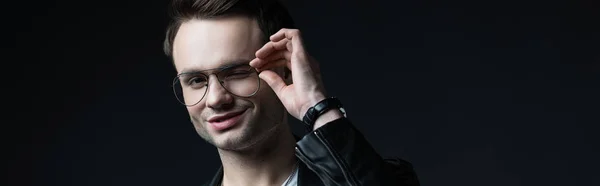 Smiling stylish brutal man in biker jacket touching eyeglasses isolated on black, panoramic shot — Stock Photo