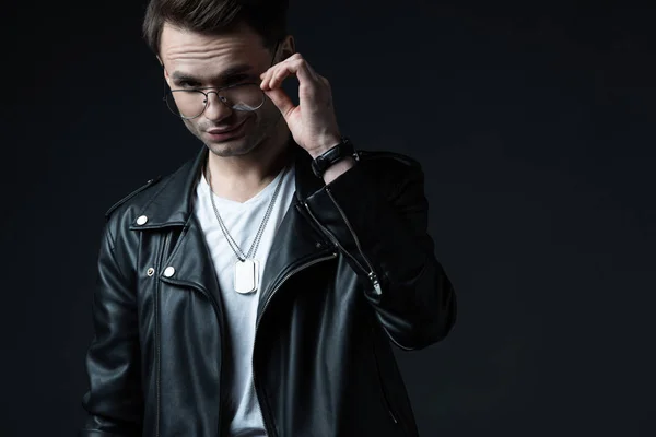 Stylish brutal man in biker jacket touching eyeglasses isolated on black — Stock Photo