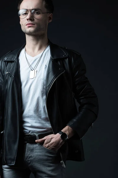 Elegante hombre brutal en chaqueta de ciclista con la mano en el bolsillo aislado en negro - foto de stock