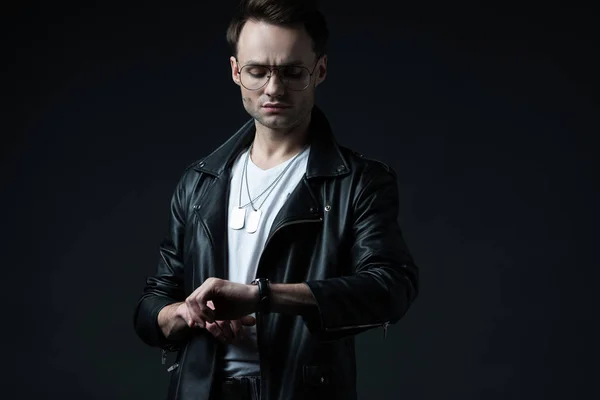 Stylish brutal man in biker jacket looking at wristwatch isolated on black — Stock Photo