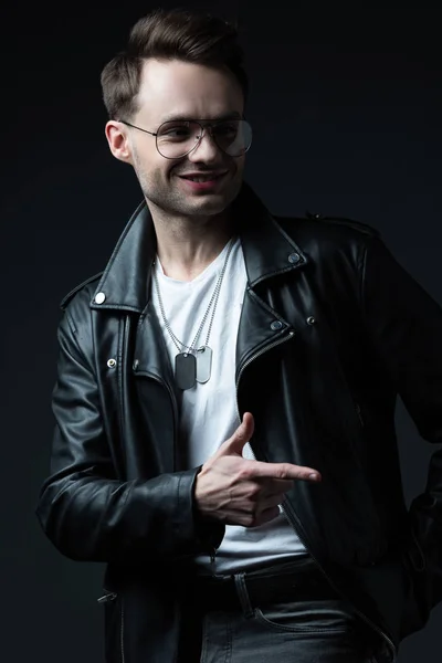 Smiling stylish brutal man in biker jacket pointing with finger aside isolated on black — Stock Photo