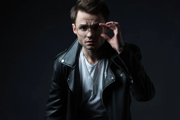 Concentrated stylish brutal man in biker jacket touching eyeglasses and looking at camera isolated on black — Stock Photo
