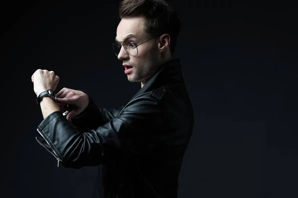 Side view of shocked stylish brutal man in biker jacket pointing with finger at wristwatch isolated on black — Stock Photo