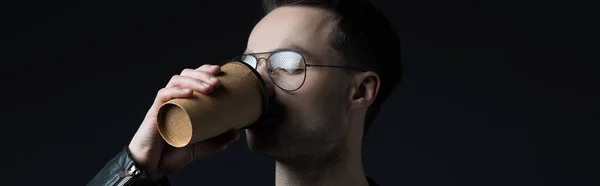 Стильный жестокий человек в очках пьет кофе из бумажной чашки, изолированной на черном, панорамный снимок — стоковое фото