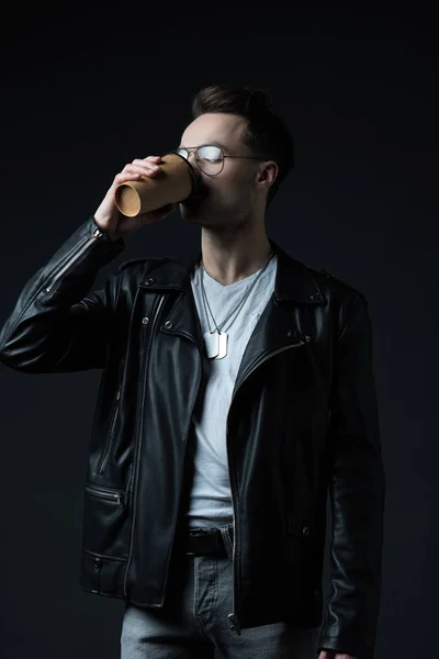 Hombre brutal con estilo en chaqueta de ciclista beber café de la taza de papel aislado en negro - foto de stock