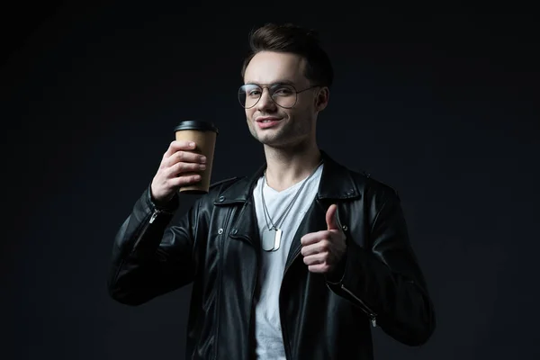 Lächelnder brutaler Mann in Bikerjacke, Kaffee to go in der Hand und Daumen hoch auf schwarz — Stockfoto