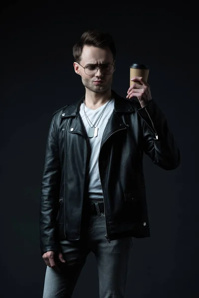 Skeptical stylish brutal man in biker jacket holding coffee to go isolated on black — Stock Photo