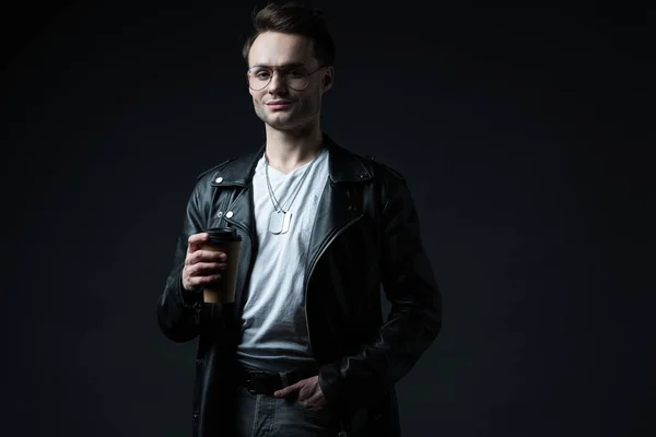 Smiling stylish brutal man in biker jacket with hand in pocket holding coffee to go isolated on black — Stock Photo